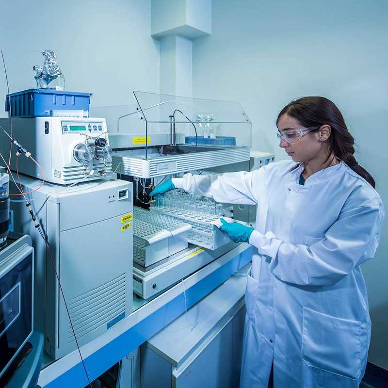 A female scientist wearing a lab coat is manipulating some glass tubes close to an HPLC system