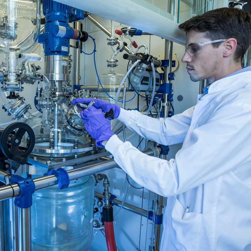 A man in glasses, lab coat, and purple gloves works on a reactor inside a fume hood.
