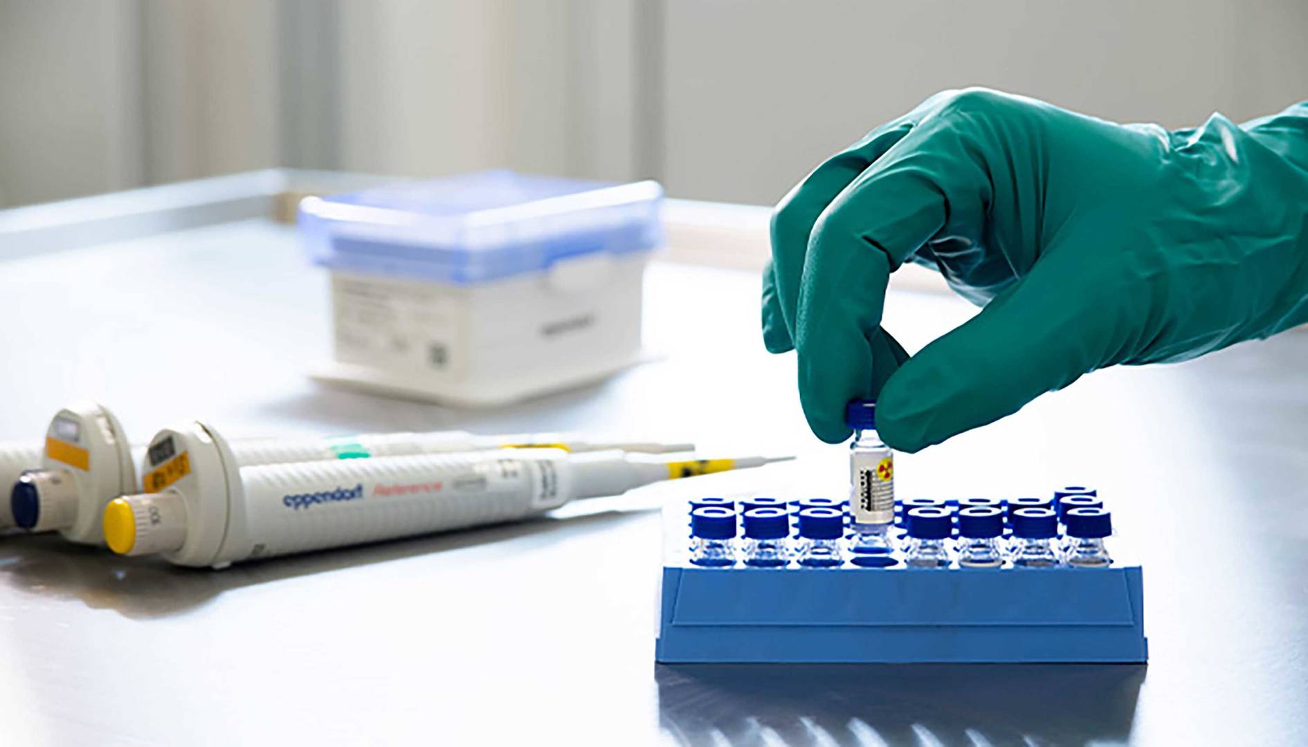 A gloved hand handles a small vial over a blue sample rack in a laboratory, with pipettes and containers in the background