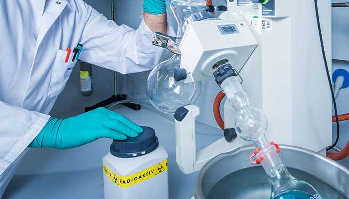 Scientist using a distillation apparatus with round bottom flasks, radioactive compound visible on the bench nearby. 